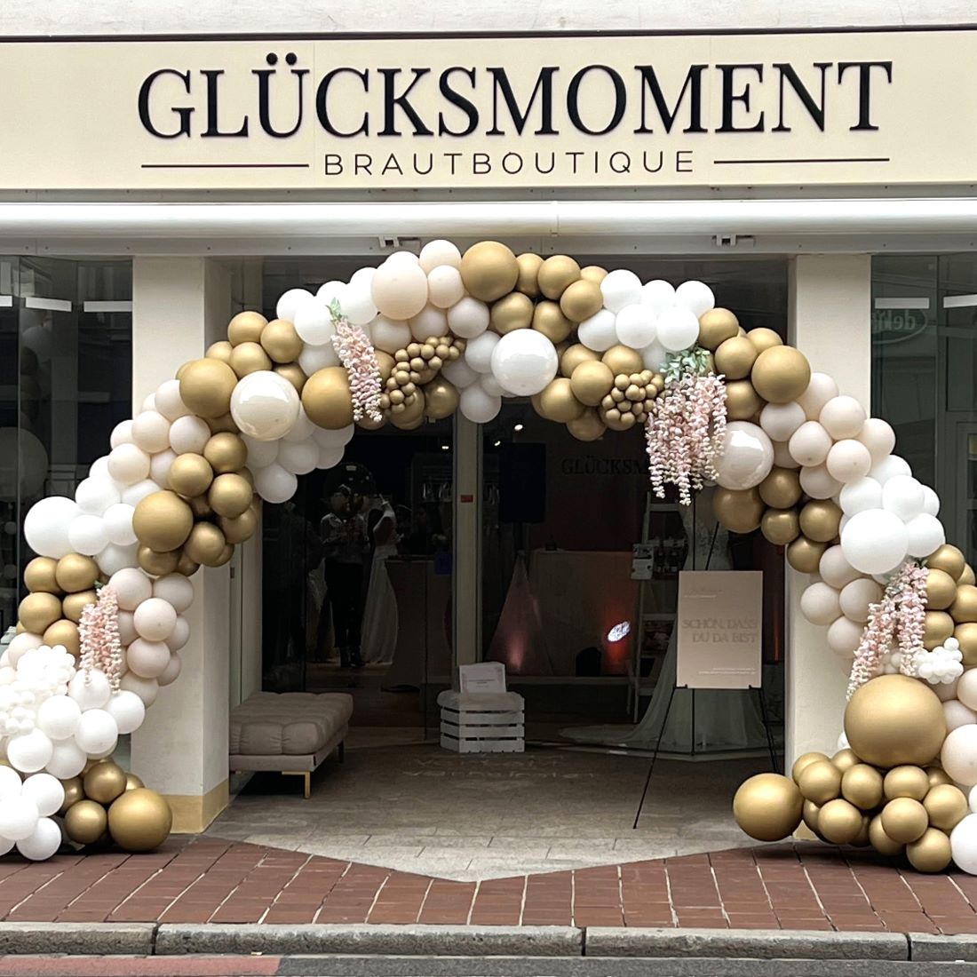 Ballonbogen in Weiß und Gold mit floralem Akzent vor dem Shop "Glücksmoment"