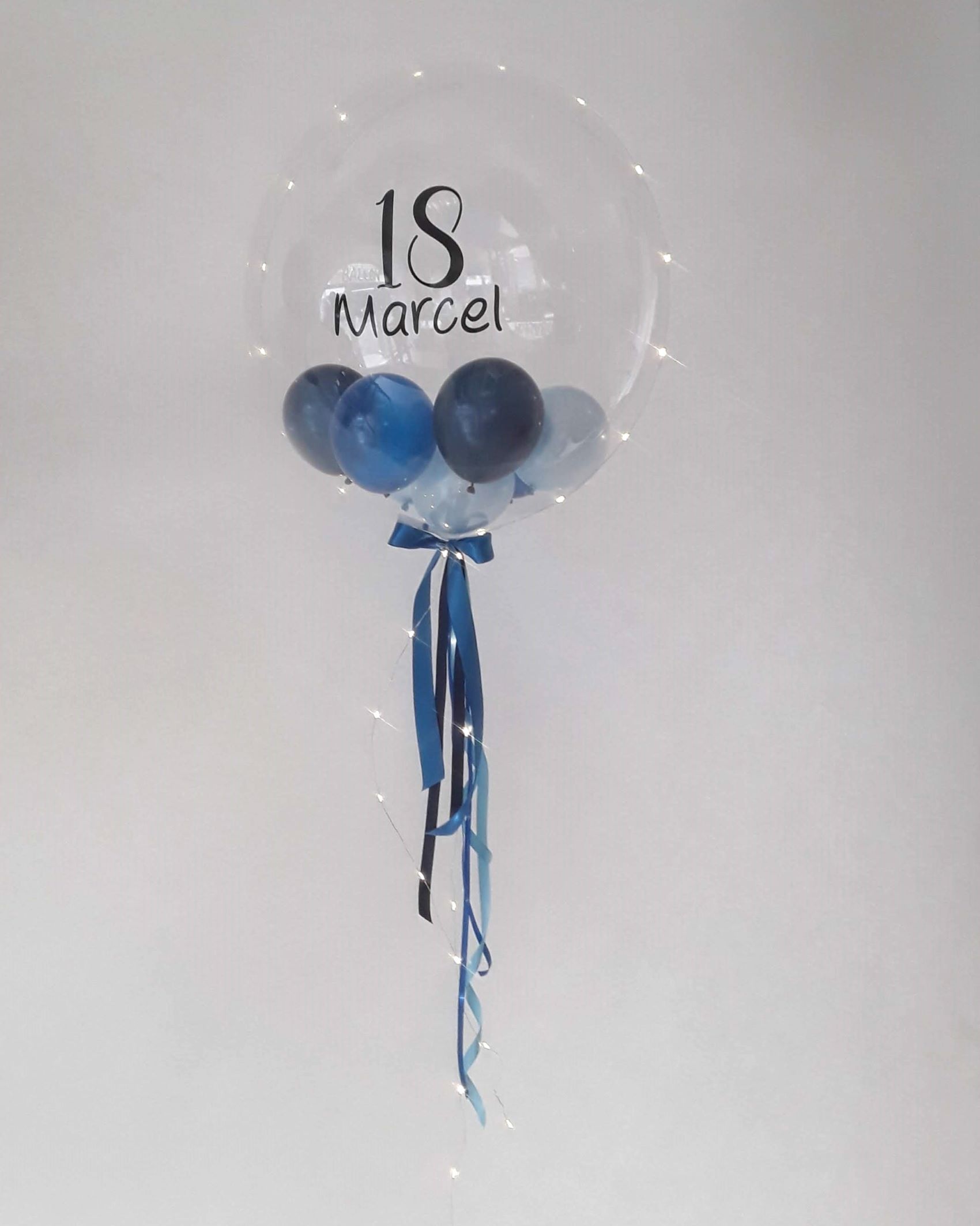 Transparenter Bubble-Ballon mit der schwarzen Aufschrift „18 Marcel“, gefüllt mit blauen und silbernen Mini-Ballons, verziert mit blauen Satinbändern und einer integrierten Lichterkette.