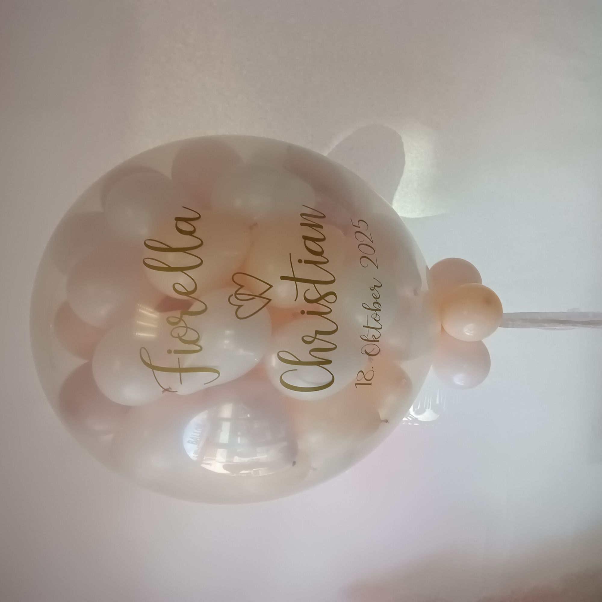 Transparenter ca.80‑cm‑Ballon mit bis zu 80 Innenballons in weiß und creme; auf dem Außenballon steht in goldener Schrift  die Namen des Brautpaars und das Datum, weißer Hintergrund