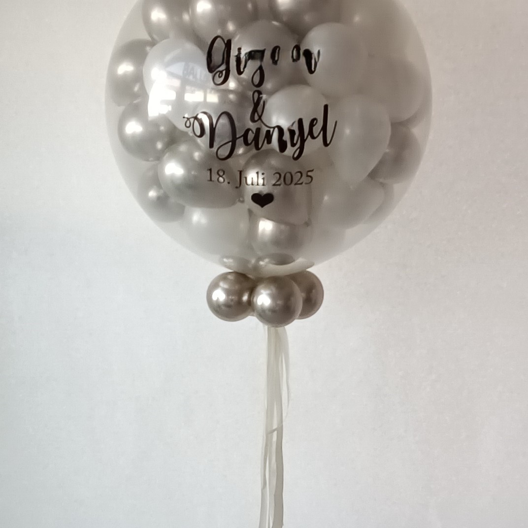 Transparenter ca.80‑cm‑Ballon mit bis zu 80 Innenballons in weiß und Weißgold; auf dem Außenballon steht in schwarzer Schrift  die namen des Brautpaars, weißer Hintergrund