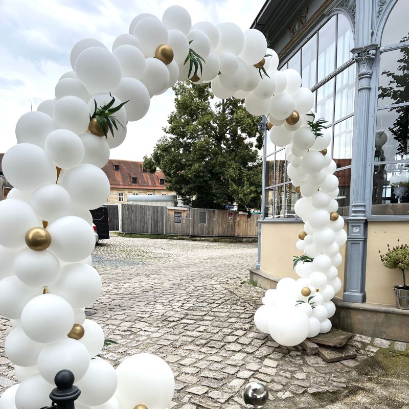 Eleganter, komplett weißer Ballonbogen mit goldenen Akzentballons und grünen Blättern, aufgebaut vor einem verglasten Gebäude