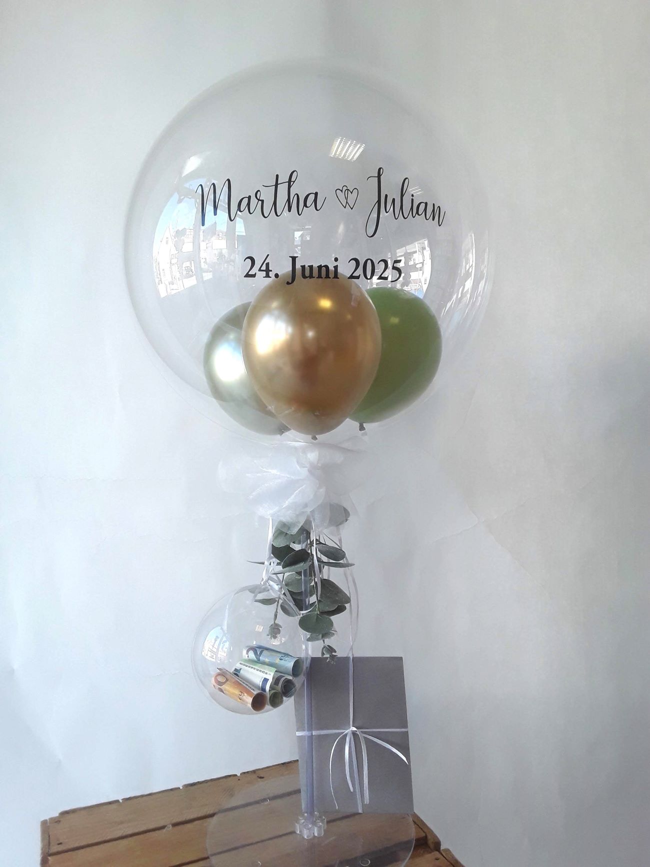 Transparenter Ballon mit Aufdruck 'Martha & Julian 24. Juni 2025' und goldenen und grünen Miniballons darin – auf Ständer mit transparentem Mini-Ballon inkl. Geldscheine, grauer Umschlag und Eukalyptus-Deko