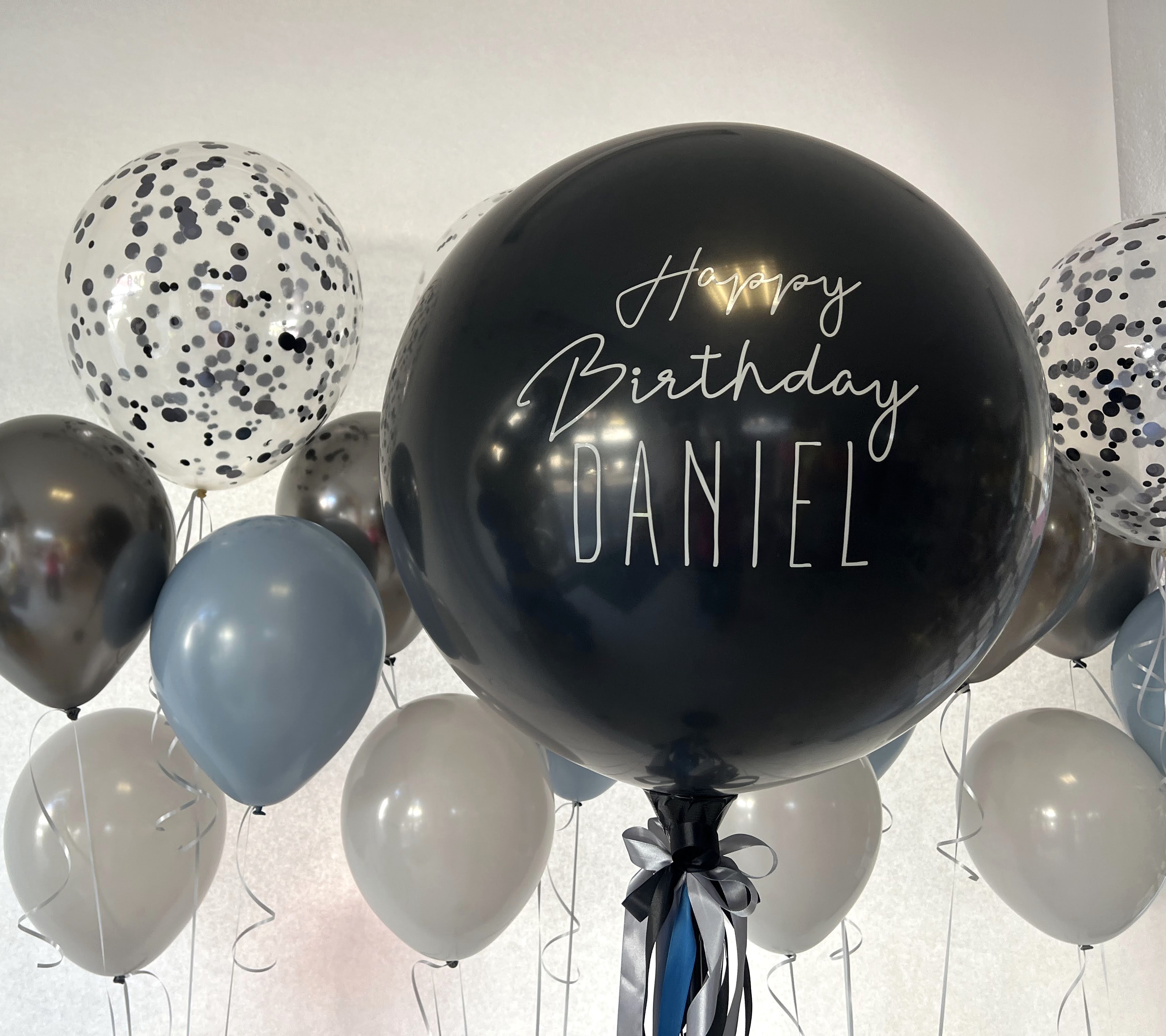 Große schwarze Ballonkugel mit weißem Aufdruck 'Happy Birthday Daniel' und dekorativer Schleife aus Satinbändern in Schwarz und Blau mit Ballons