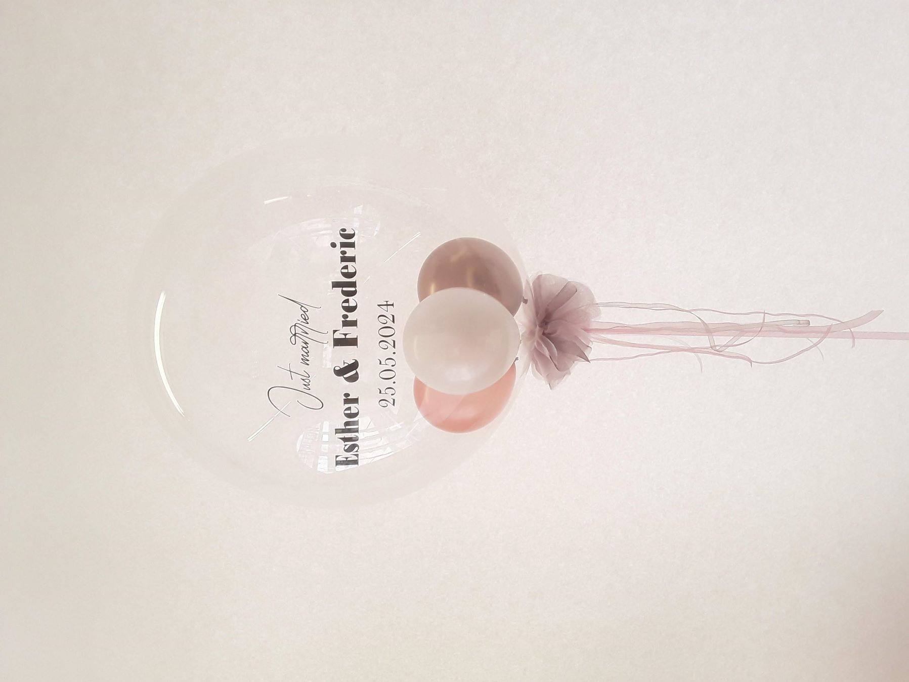 Klarer Ballon mit personalisiertem Aufdruck „Just married Esther & Frederic 25.05.2024“ und drei schwebenden Ballons in Rosa, Weiß und Kupfer sowie einer Schleife aus Tüll.