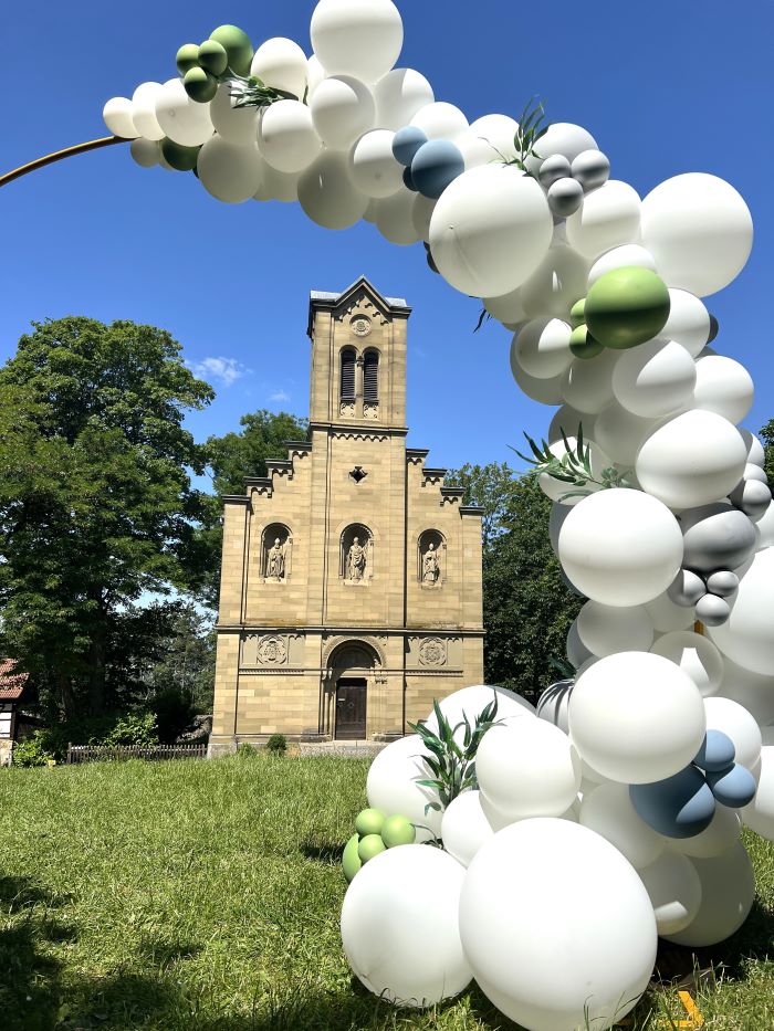 Halbkreisförmiger Ballonbogen in Weiß mit grünen und blauen Akzenten, dekoriert mit Eukalyptuszweigen und Ballon mit Schriftzug „Schön, dass ihr da seid!“2