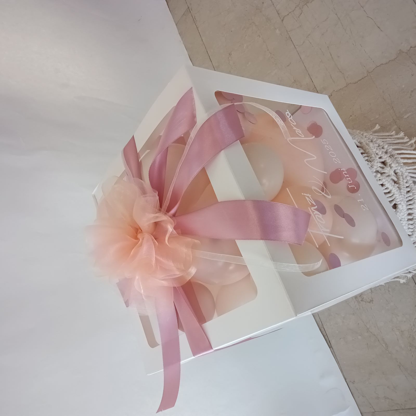 Weiße Ballonbox mit apricot- und roséfarbenen Luftballons, Konfetti, transparenter Front, rosa Satinband und Tüllschleife.