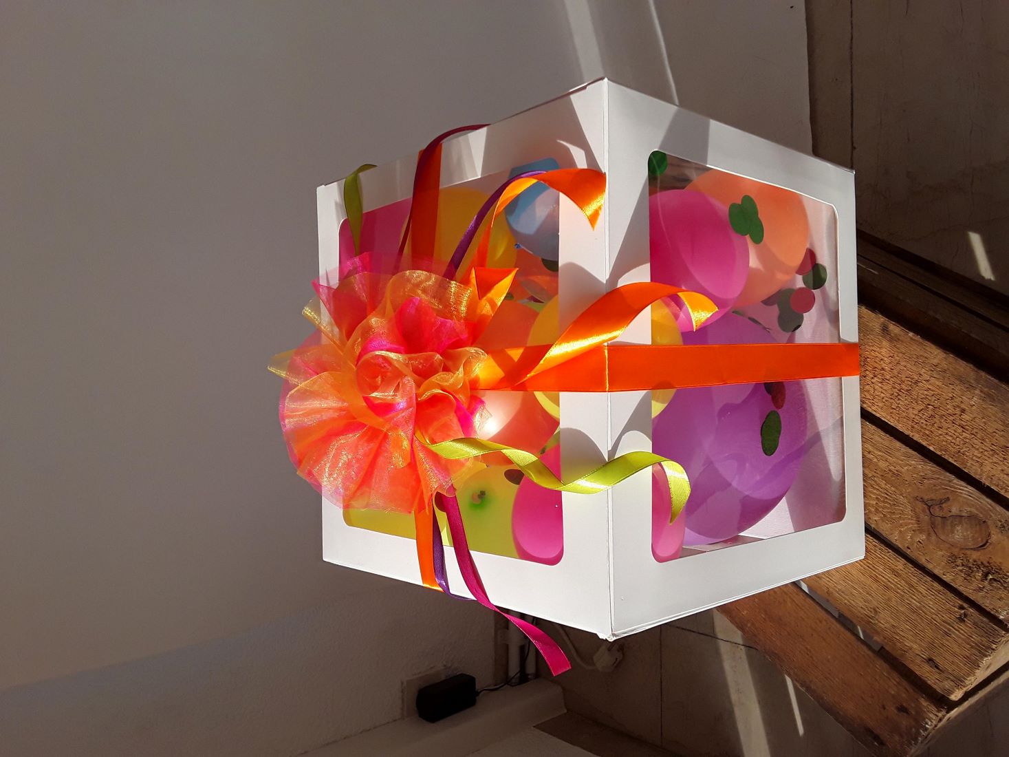 Weiße Ballonbox mit Ballons in Lila, Orange, Gelb, Pink und Grün, dekoriert mit bunter Schleife und Konfetti.