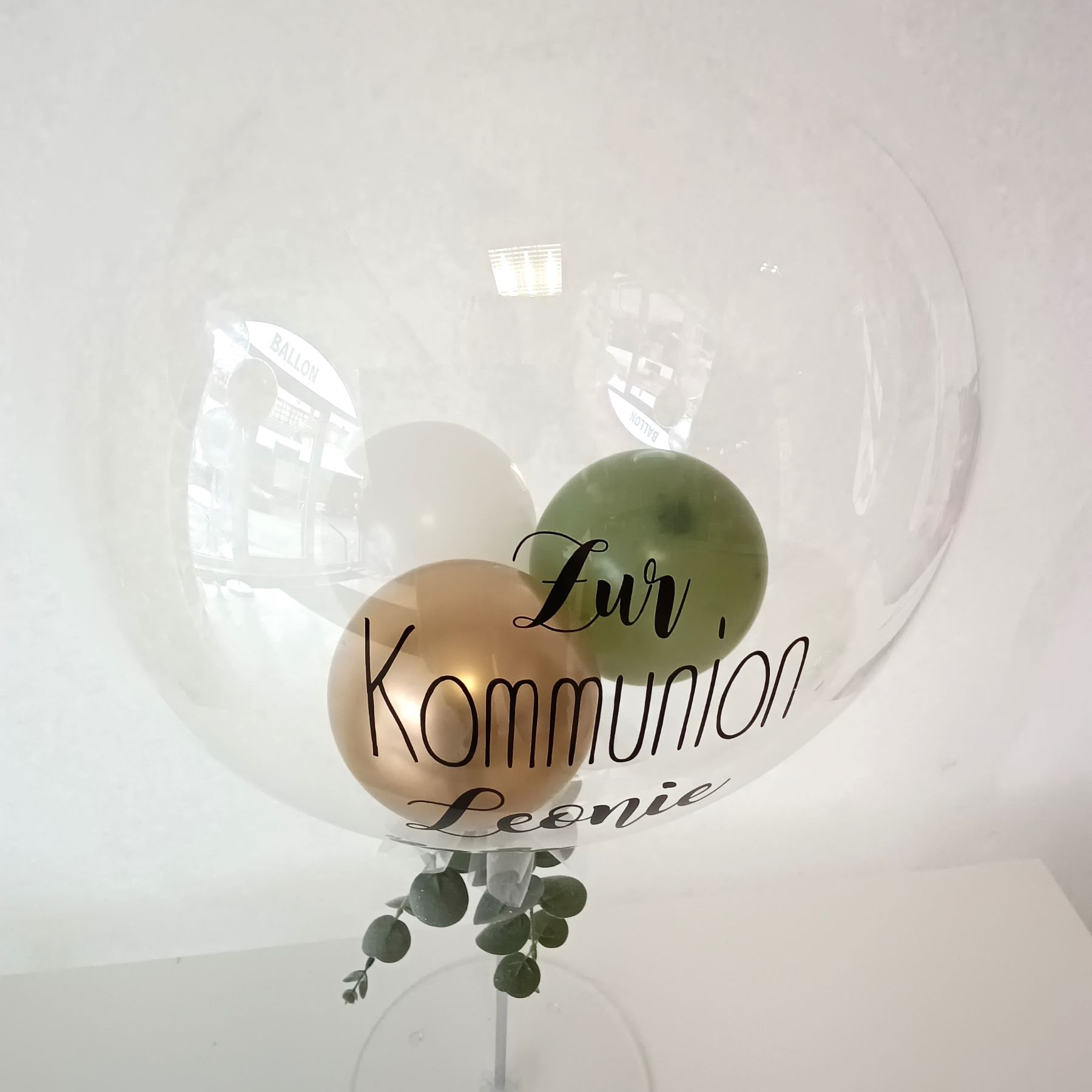 Tischständer mit transparentem Aqua-Ballon gefüllt mit  3 kleinen Ballons in grün, gold und weiß. Aufdruck  mit "zur Kommunion Leonie". Eukalyptusdeko und weiße Schleife