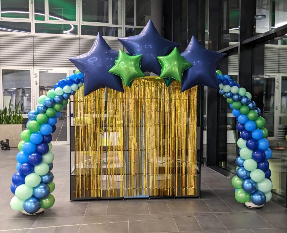 Ballonbogen aus blauen, grünen und mintfarbenen Latex- und Metallic-Ballons mit fünf Stern-Folienballons in Blau und Grün mit goldenem Vorhang in einer Halle