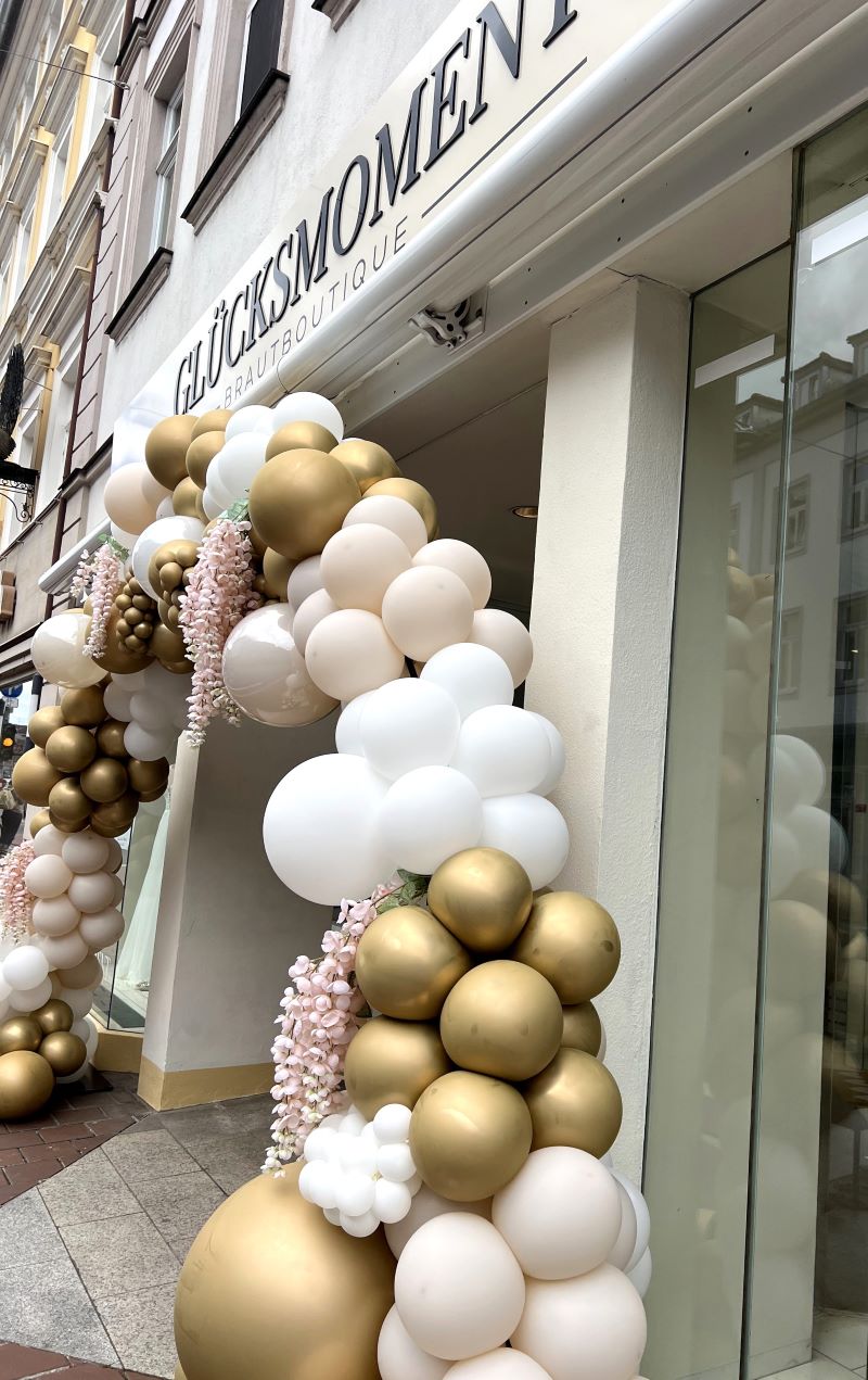 Ballonbogen in Weiß und Gold mit floralem Akzent vor dem Shop "Glücksmoment"