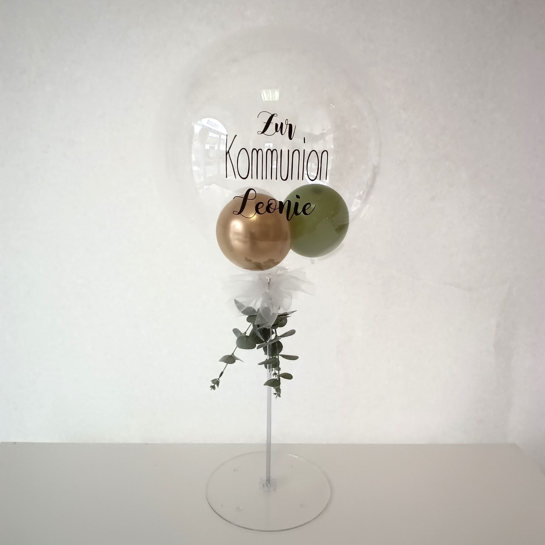 Tischständer mit transparentem Aqua-Ballon gefüllt mit  3 kleinen Ballons in grün, gold und weiß. Aufdruck  mit "zur Kommunion Leonie". Eukalyptusdeko und weiße Schleife