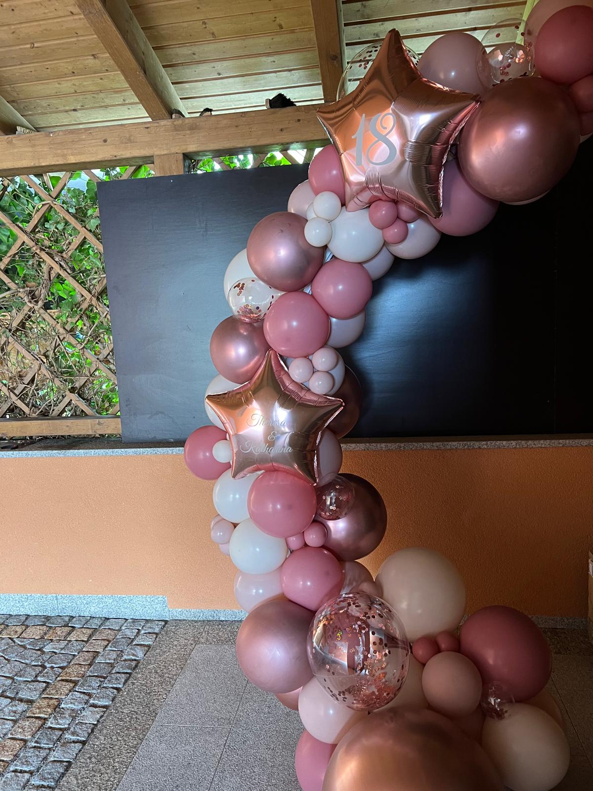 Ringbogen aus Ballons in Roségold, Rosa, Pink und Weiß, dekoriert mit großen Sternfolienballons mit Aufdruck '18' und 'Theresa&Katharina' und transparenten Konfettiballons